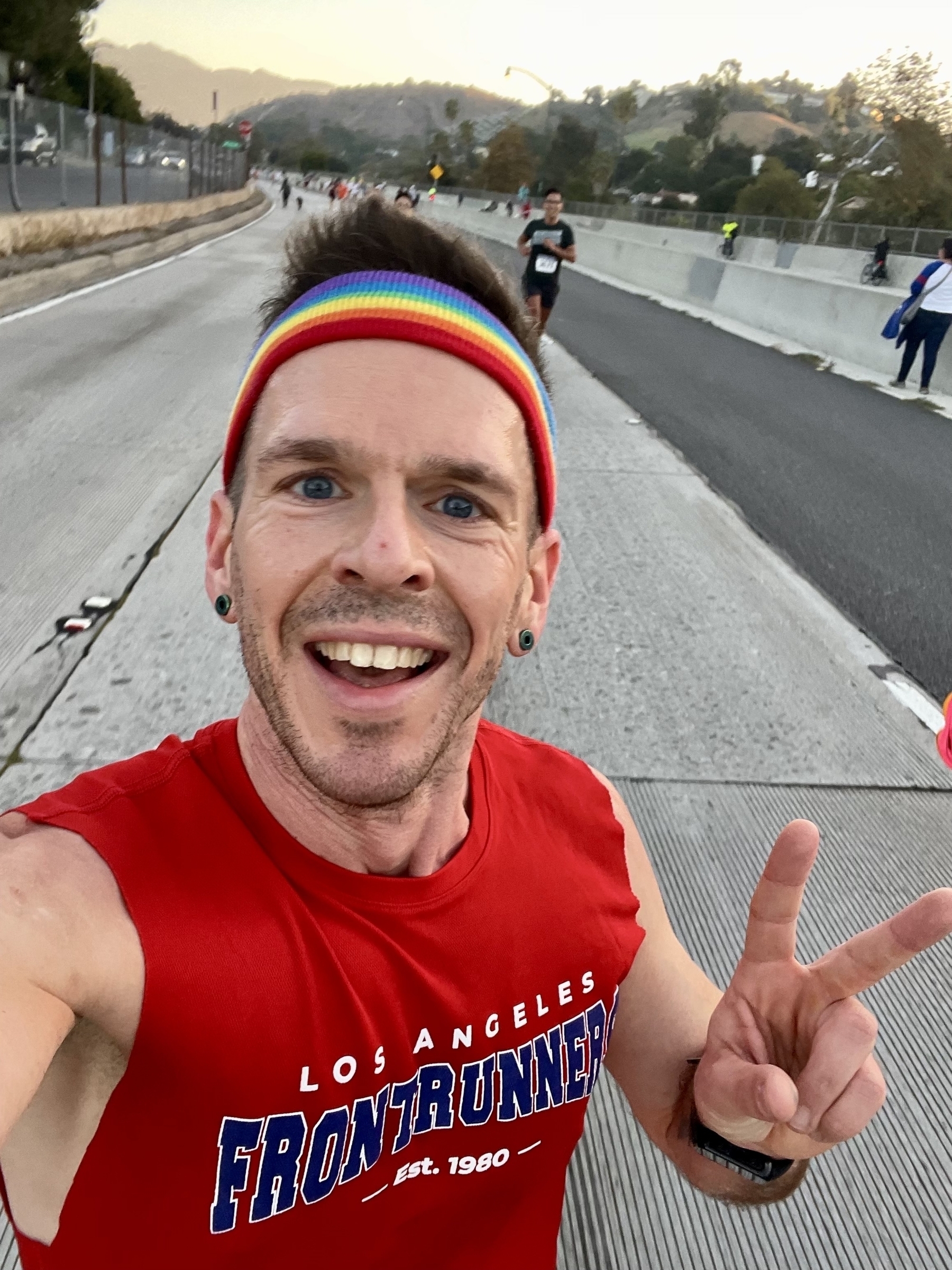 A man in a sleeveless red t-shirt and rainbow headband takes a selfie while running down a car-free highway with other runners