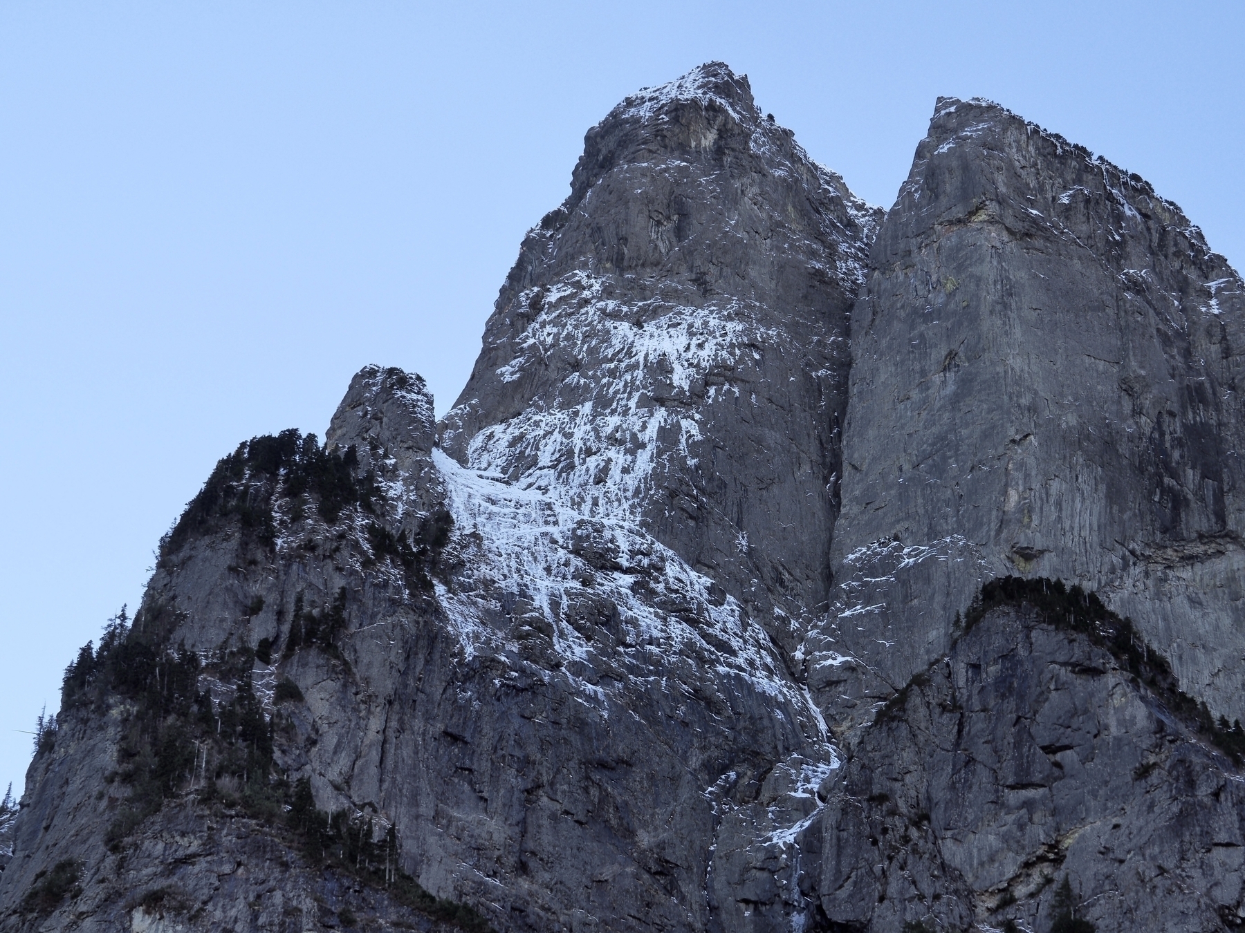 A triangular mountain peak with a steep rocky face, edged with dark trees and patches of snow
