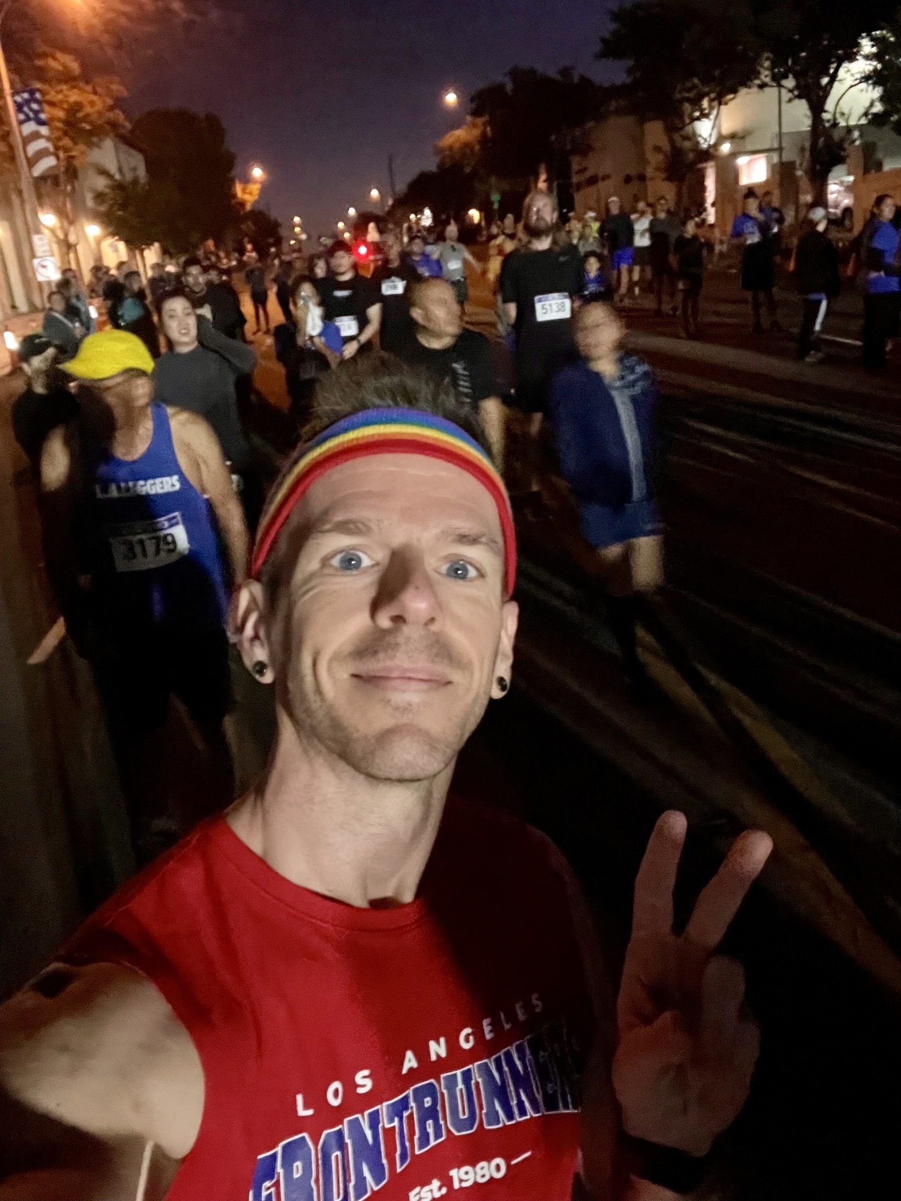 A man in a red sleeveless t-shirt and rainbow headband takes a selfie in the starting line crowd for a road race, in early morning darkness 