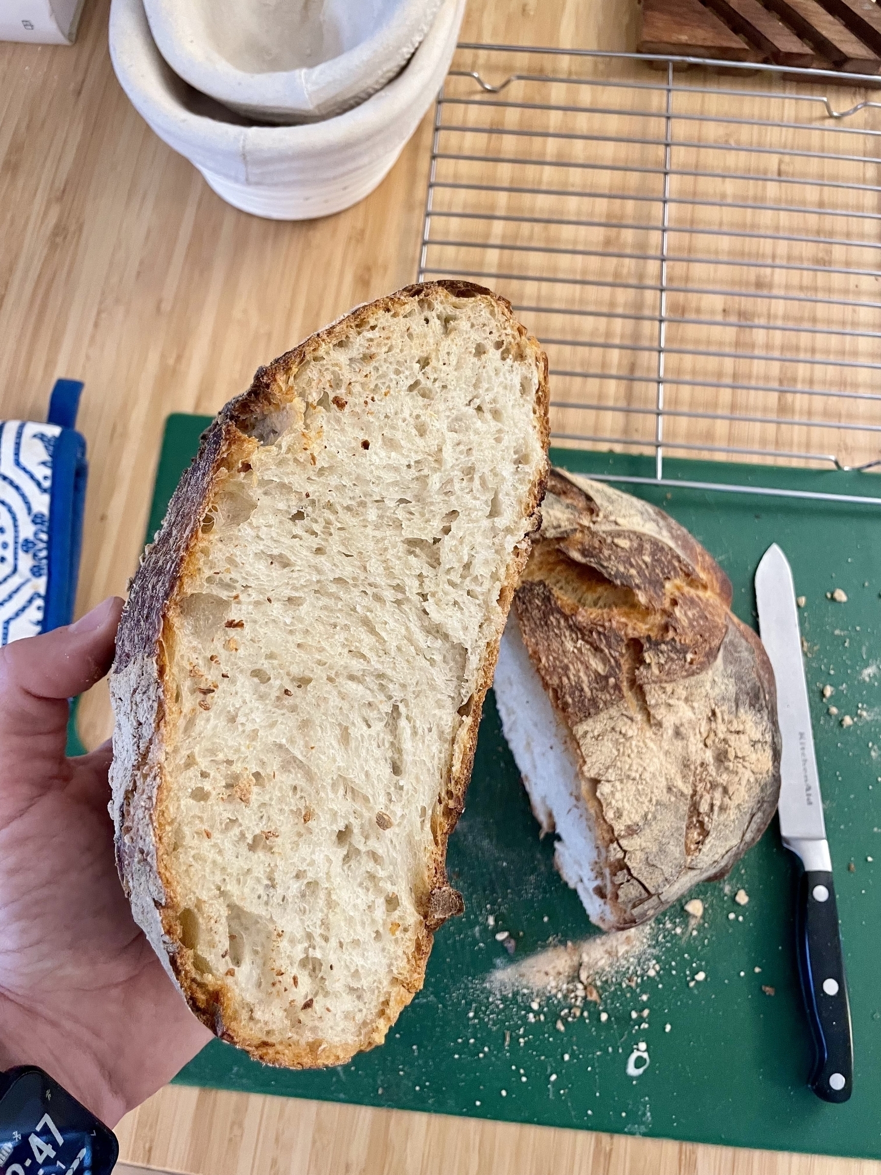 The boule cut in half, one half sitting amidst crumbs on the cutting board, the other held up to the camera to show an even crumb and thick crust