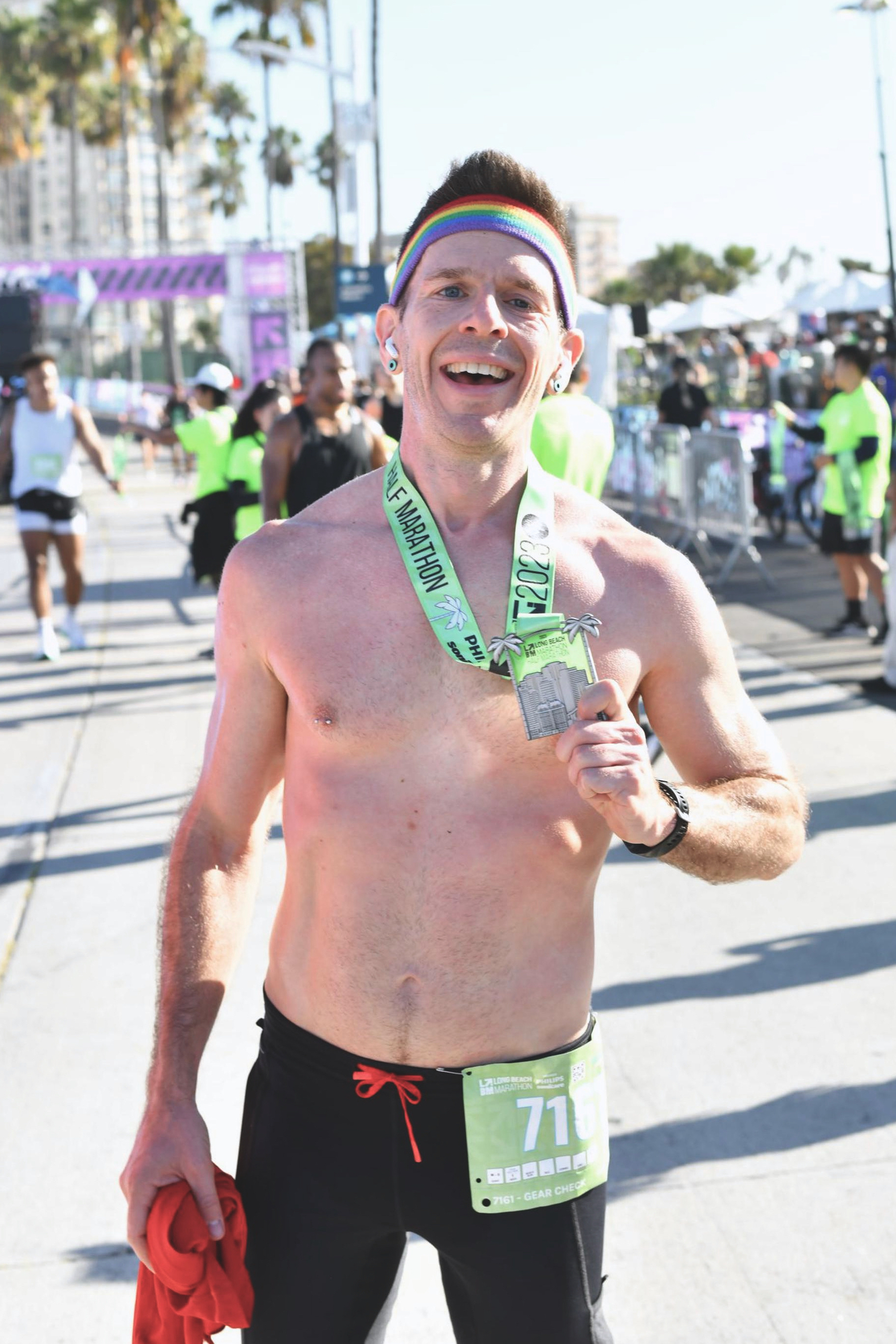A man in short tights and a rainbow headband standing in the finish area of a race, holding up a finisher medal